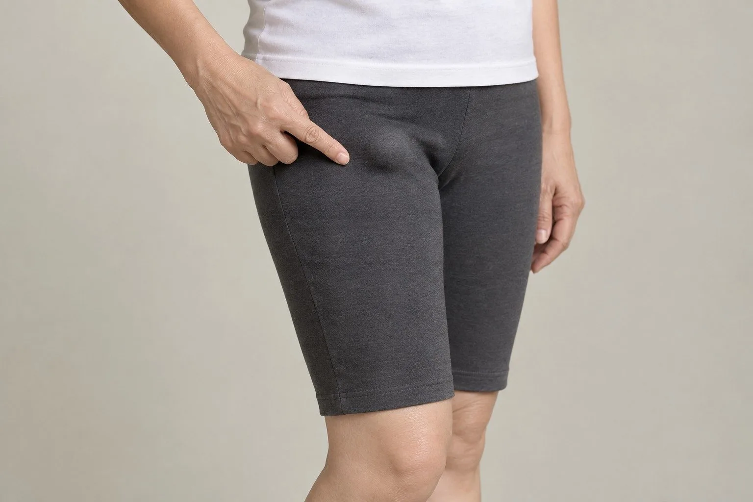 A woman in fitted grey shorts points to a small rounded swelling high on her inner thigh, just below the fold of the groin
